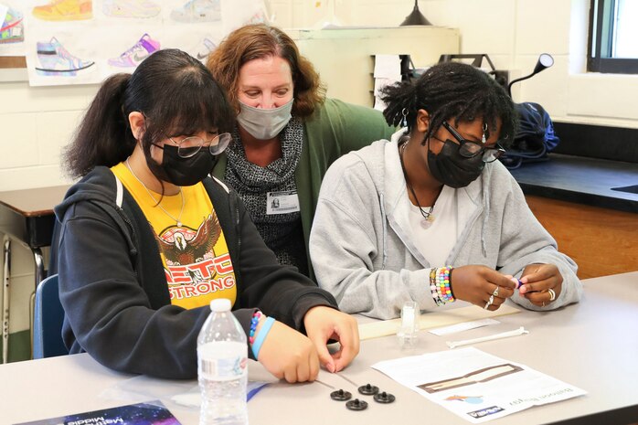 NSWC IHD Chief Technology Officer Dr. Kerry Clark sits between two students to help them figure out the best way to build their balloon buggies during their science class.