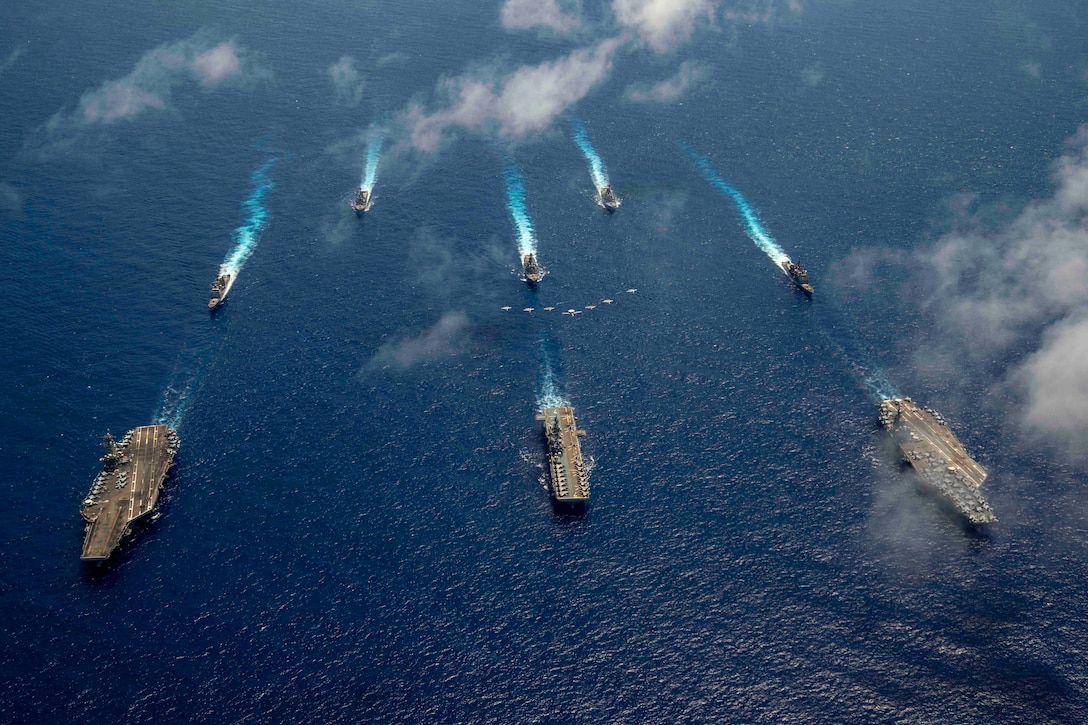 Eight ships sail in formation as Navy aircraft fly above them.