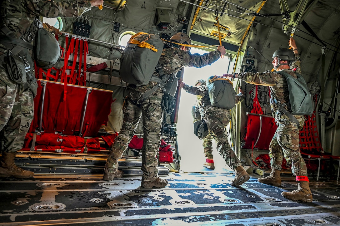 A soldier leans out an aircraft before leaping as others stand in the aircraft.
