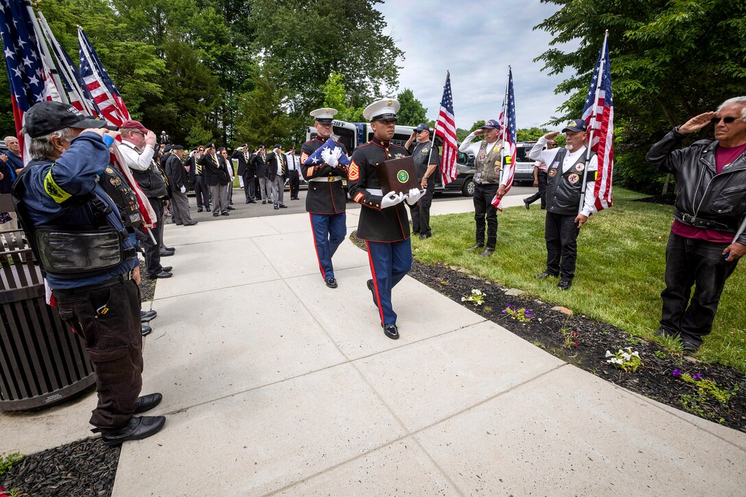 A Marine carries an urn and another carries an American flag past a group of saluting civilians.