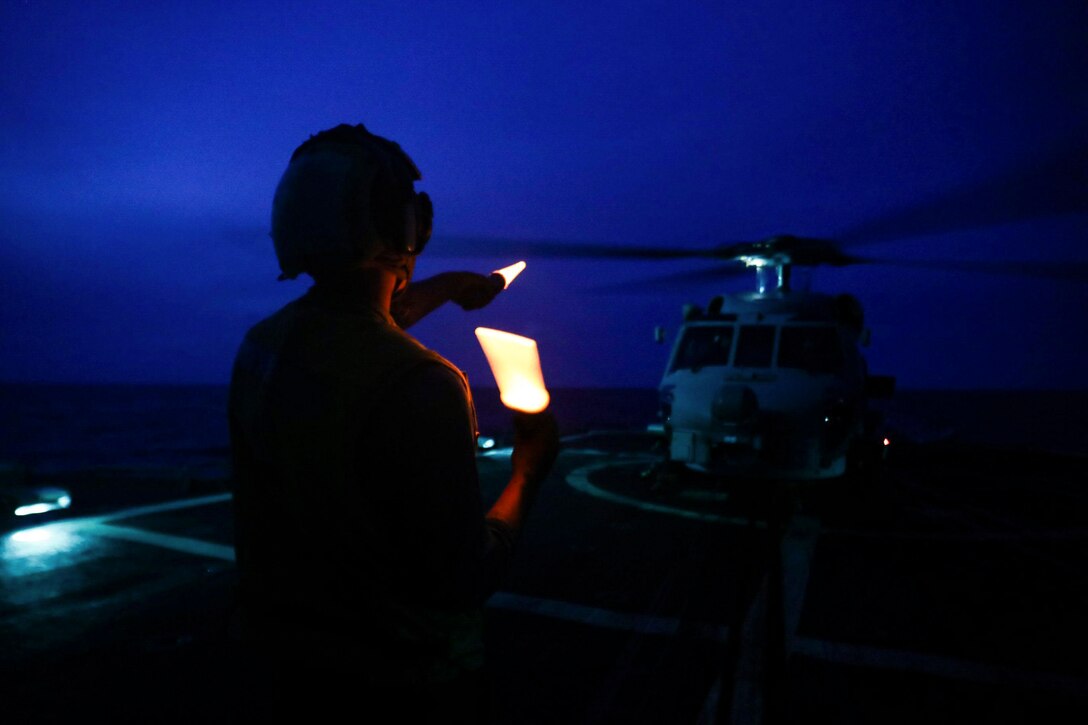 A sailor directs a helicopter aboard a ship at sea in the dark.