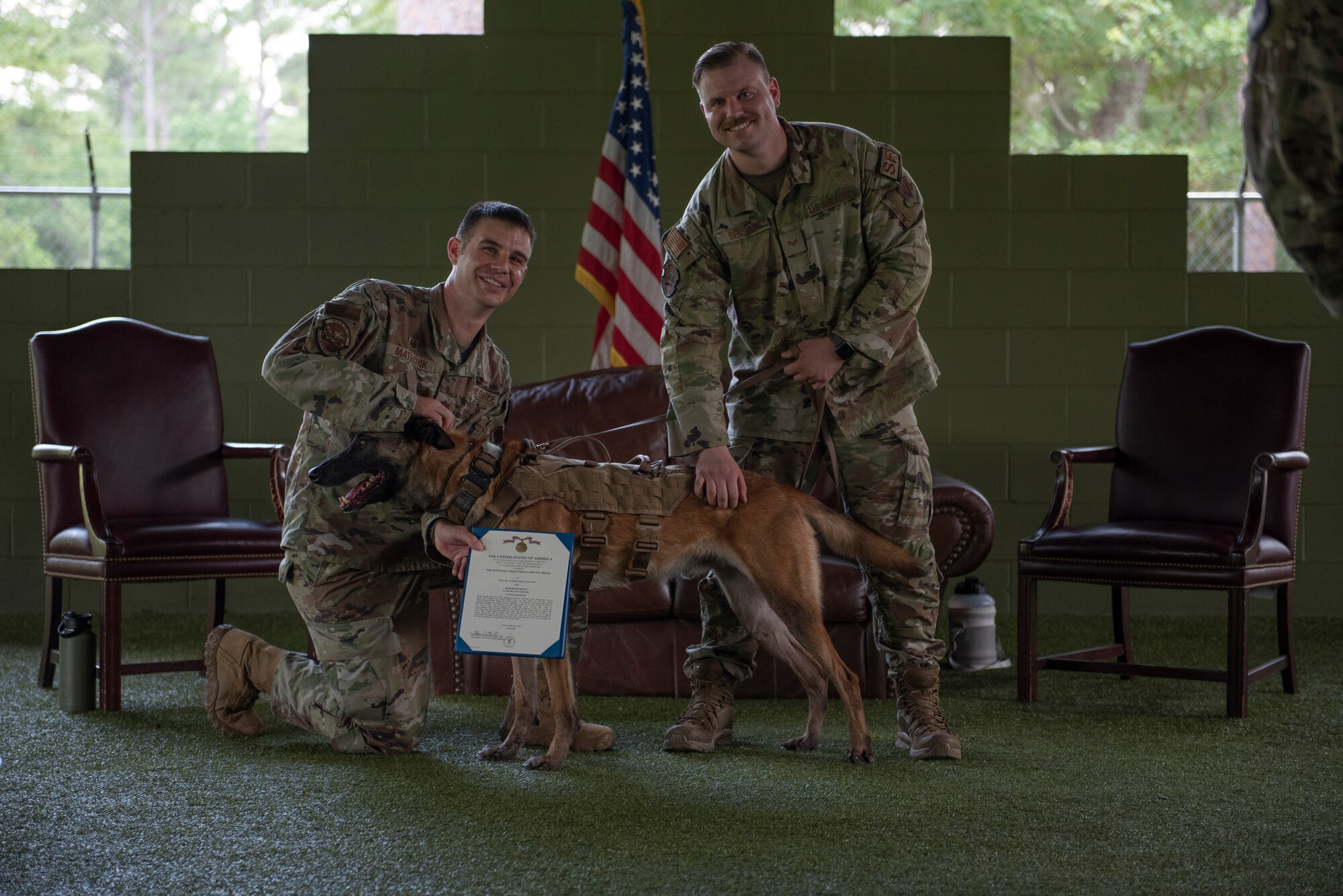 A photo two Airmen standing behind a military working dog. One Airman is holding a medal.