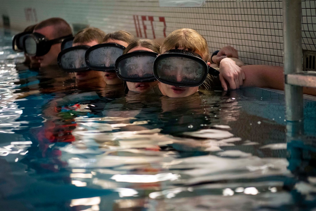 Army ROTC cadets and U.S. Air Force Academy cadets wearing goggles hang on to the wall inside of a pool.