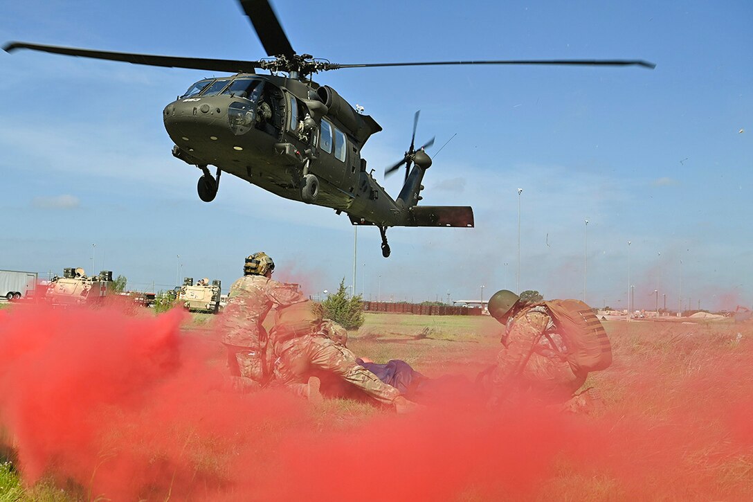 An Army helicopter flies over service members surrounded by clouds of red smoke.