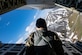 An Airman sits on the open ramp of a C-130 as it flies over mountains.