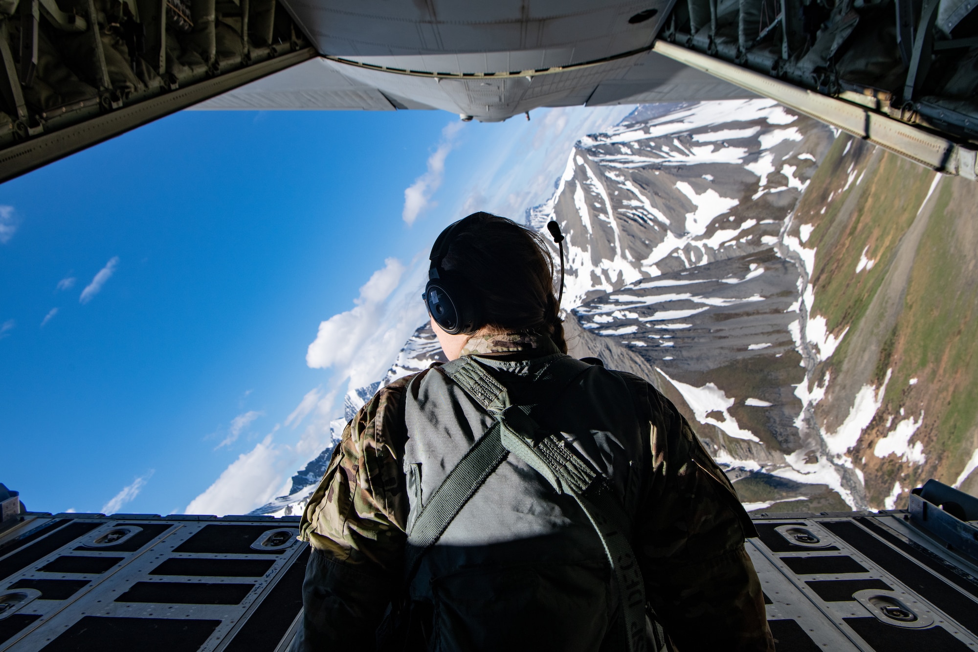 An Airman sits on the open ramp of a C-130 as it flies over mountains.