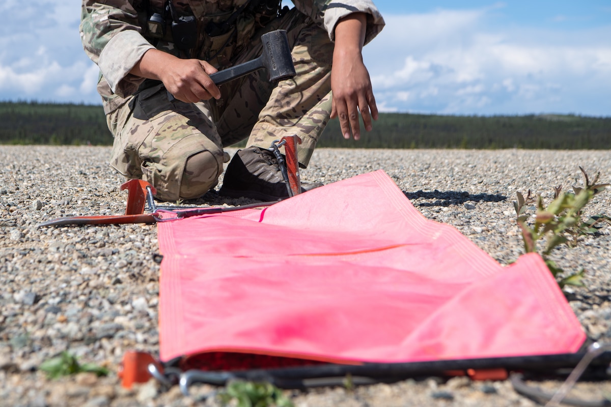 An Airmen hammers in a brightly colored banner on a dirt runway.