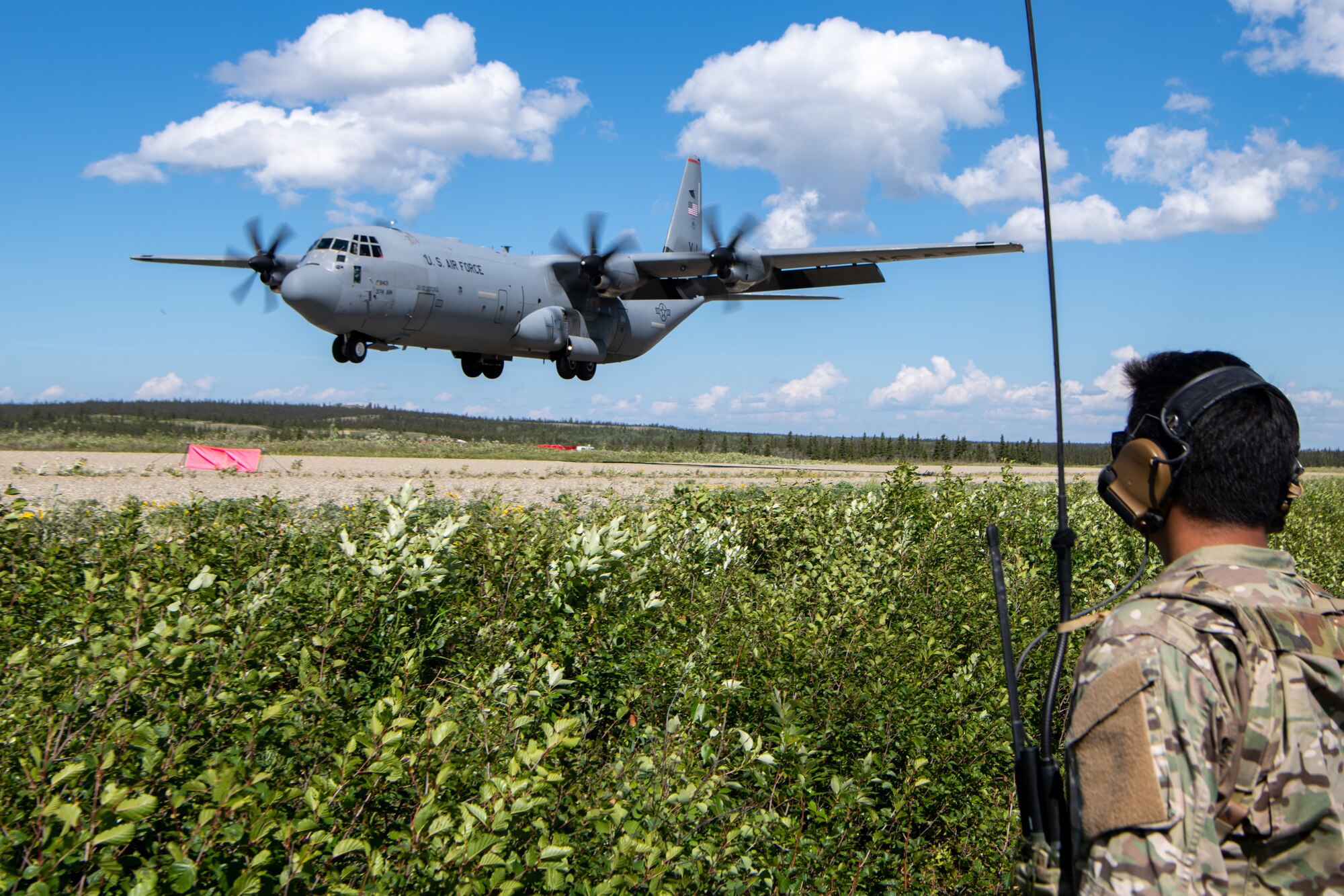 An Airmen observes a C-130 come in for a landing on a dirt runway.