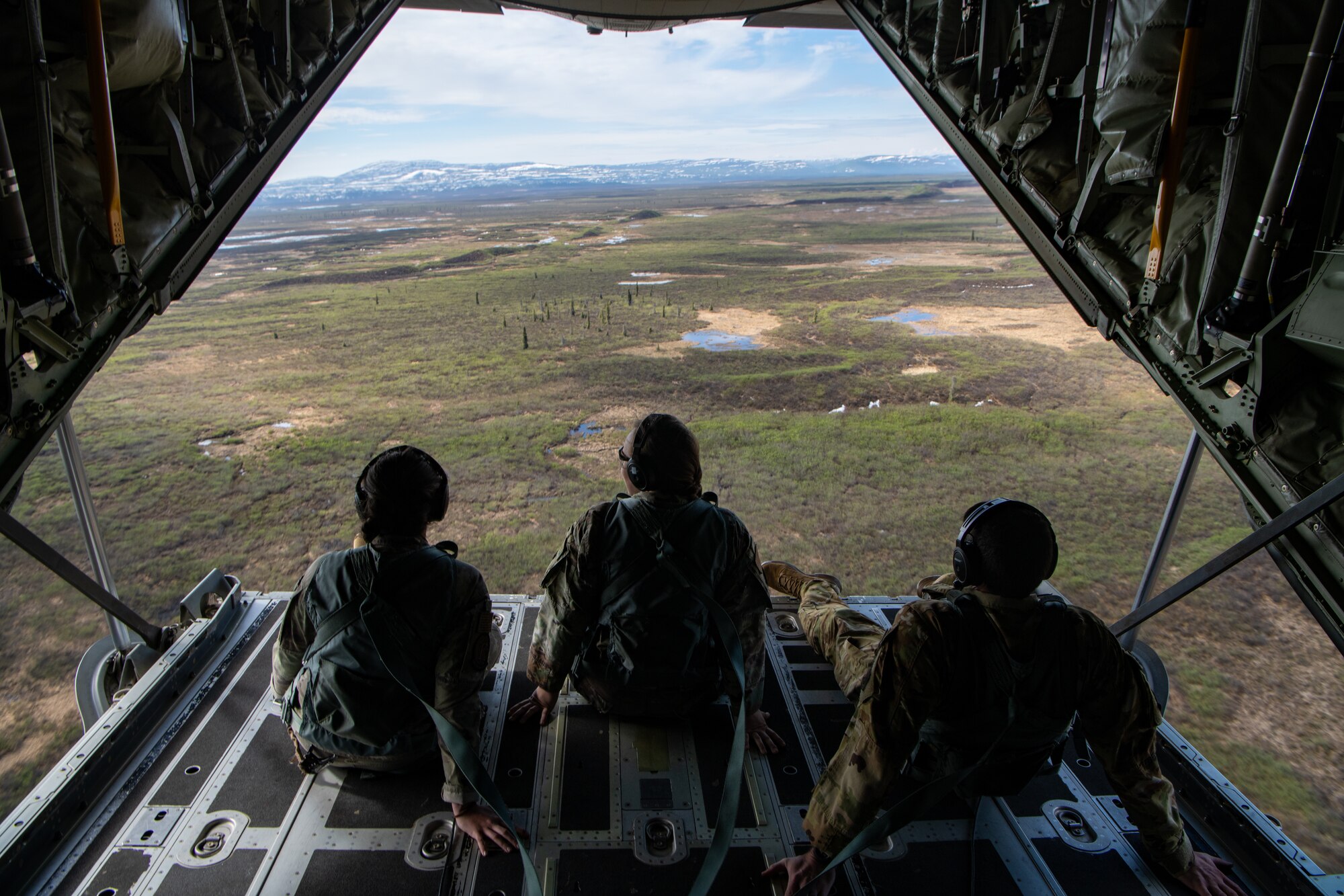 Airmen sit on the open ramp of a C-130 as it flies over mountains.