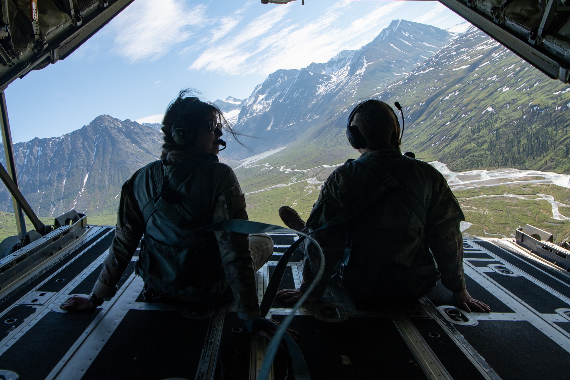 Airmen sit on the open ramp of a C-130 as it flies over mountains.