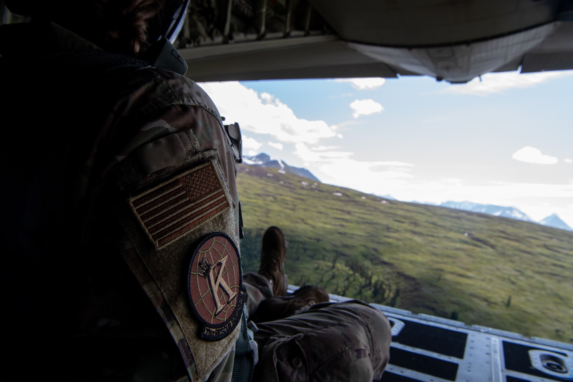 An Airman sits on the open ramp of a C-130 as it flies over mountains.