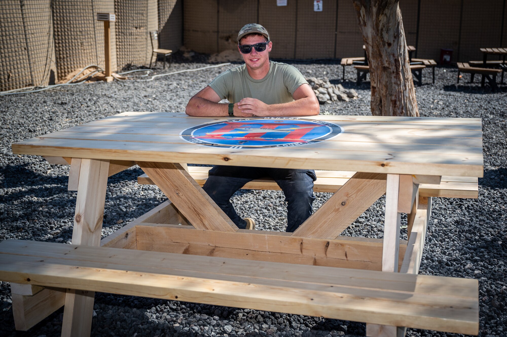 Airman First Class Bryson Doll, 332d Expeditionary Civil Engineer Squadron structural apprentice, sits at the picnic table he designed, built, and painted for the 332d Air Expeditionary Wing Top III Council at an undisclosed location in Southwest Asia, May 18, 2022. During his deployment, Doll has volunteered 50 hours, before and after work, on various woodworking projects that include bag toss boards, a bench crafted from a shipping palate, a picnic table, a tail fin coin holder, and several other projects. (U.S. Air Force photo by Master Sgt. Christopher Parr)