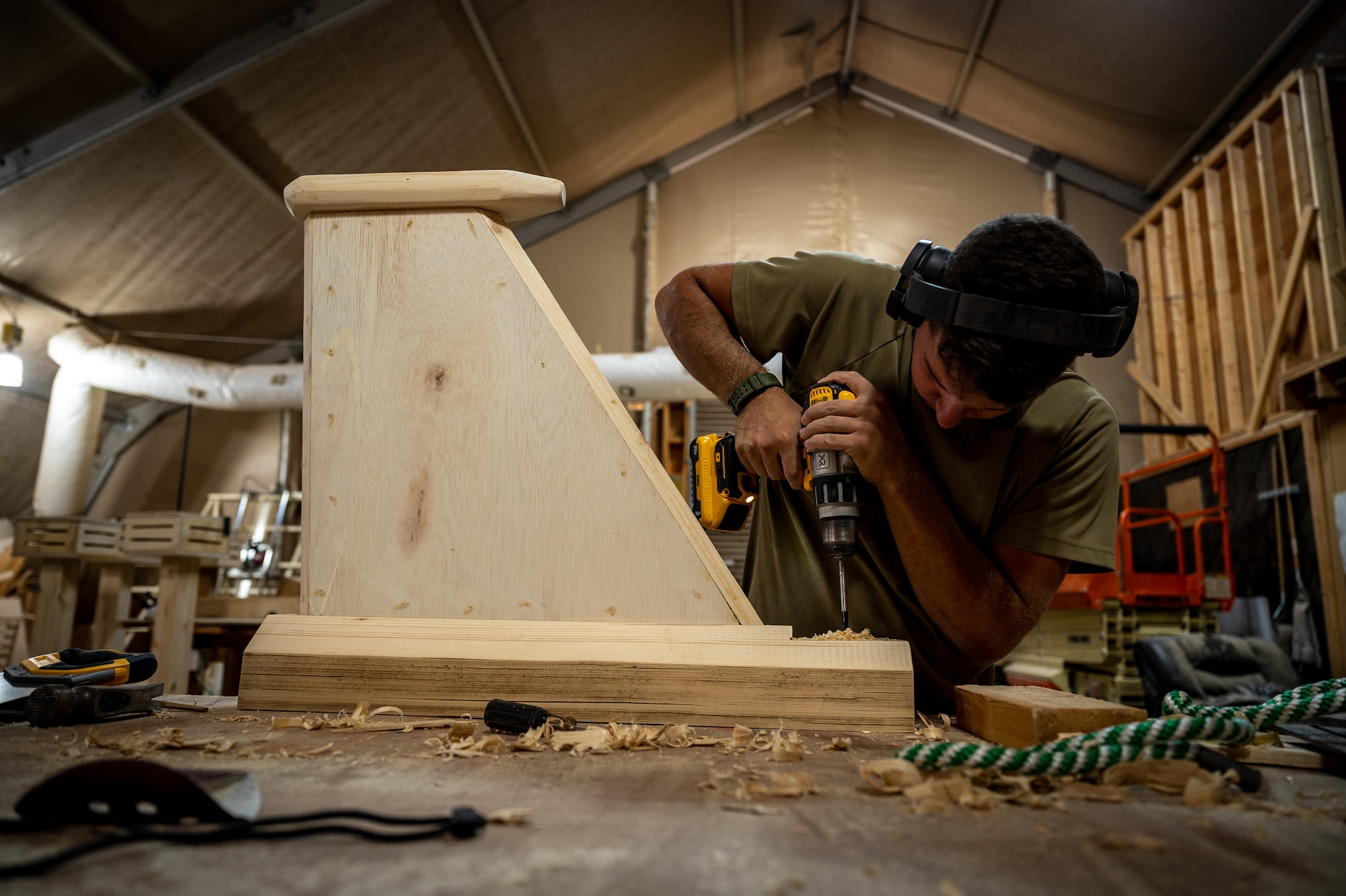 Airman First Class Bryson Doll, 332d Civil Engineer Squadron structural apprentice, drills into the base of a woodworking project at an undisclosed location in Southwest Asia, May 21, 2022. During his deployment, Doll has volunteered 50 hours, before and after work, on various woodworking projects that include bag toss boards, a bench crafted from a shipping palate, a picnic table, a tail fin coin holder, and several other projects. (U.S. Air Force photo by Master Sgt. Christopher Parr)