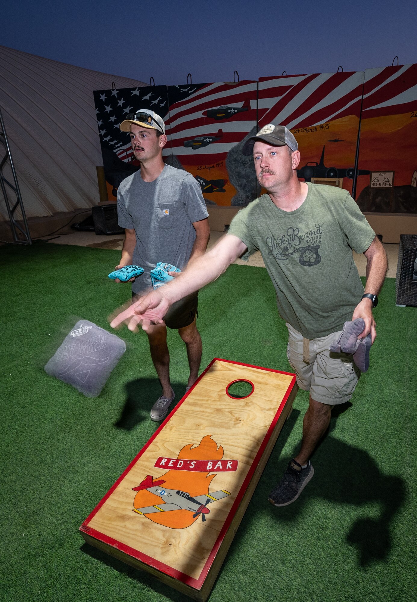 Master Sgt. Timothy Travis, 332 Expeditionary Civil Engineer Squadron operations management superintendent, tossing the bag, and Senior Airman Joshua Grimm, 332 ECES structural journeyman, warm up for a bag toss tournament at an undisclosed location in Southwest Asia, June 9, 2022. Airman First Class Bryson Doll, 332d Expeditionary Civil Engineer Squadron structural apprentice, used his woodworking skills and free time to build and hand paint the bag toss boards that provide morale opportunities for Red Tails. (U.S. Air Force photo by Master Sgt. Christopher Parr)