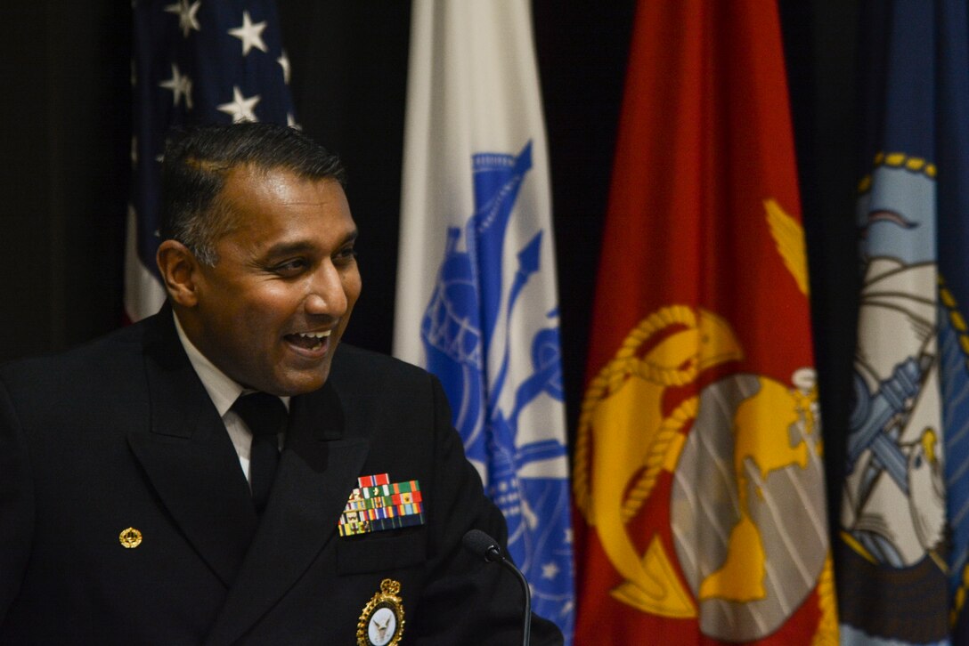 A man in a US Navy uniform speaks at a podium in an auditorium.