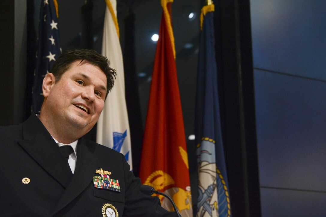 A man in a US Navy uniform speaks at a podium in an auditorium.