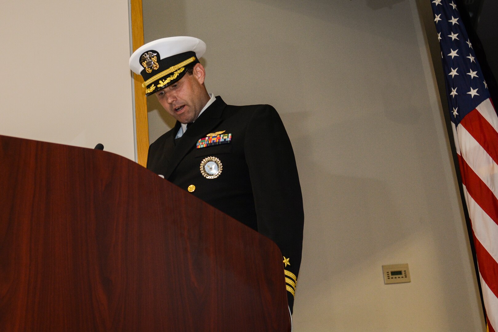 A man in a US Navy uniform speaks at a podium in an auditorium.