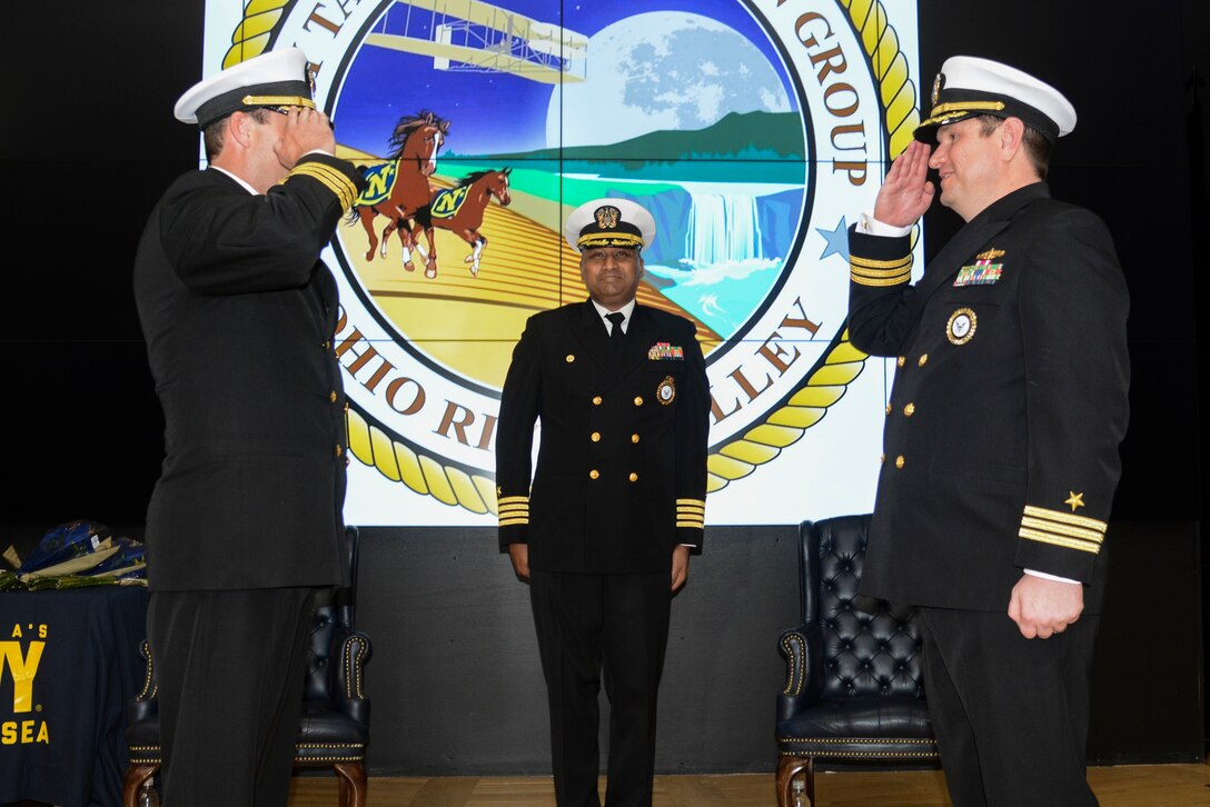 Men in US Navy uniforms conduct a change of command ceremony on stage in an auditorium.