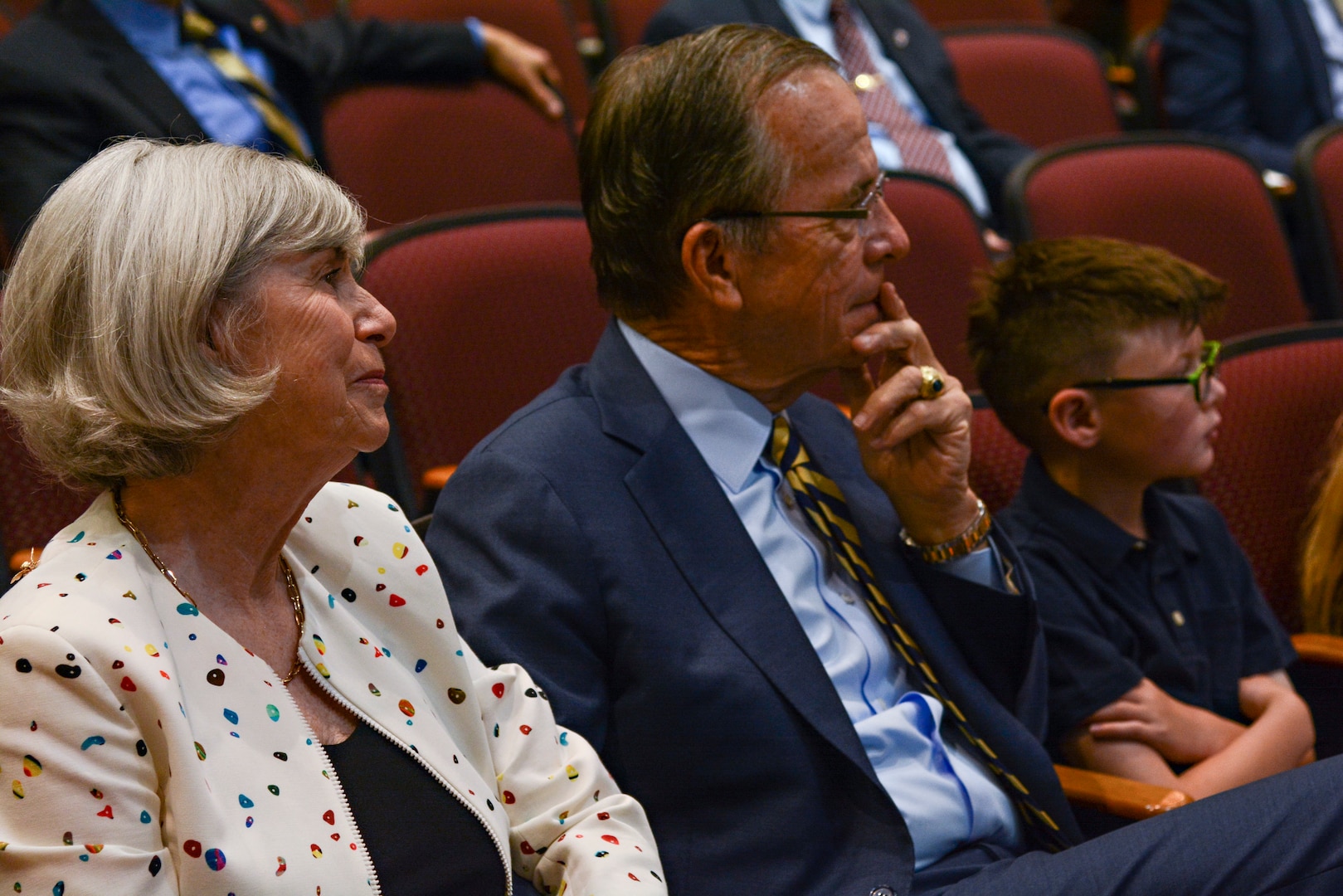A man in a suit sits next to a woman in a white dress with a boy with glasses next to them in an auditorium.