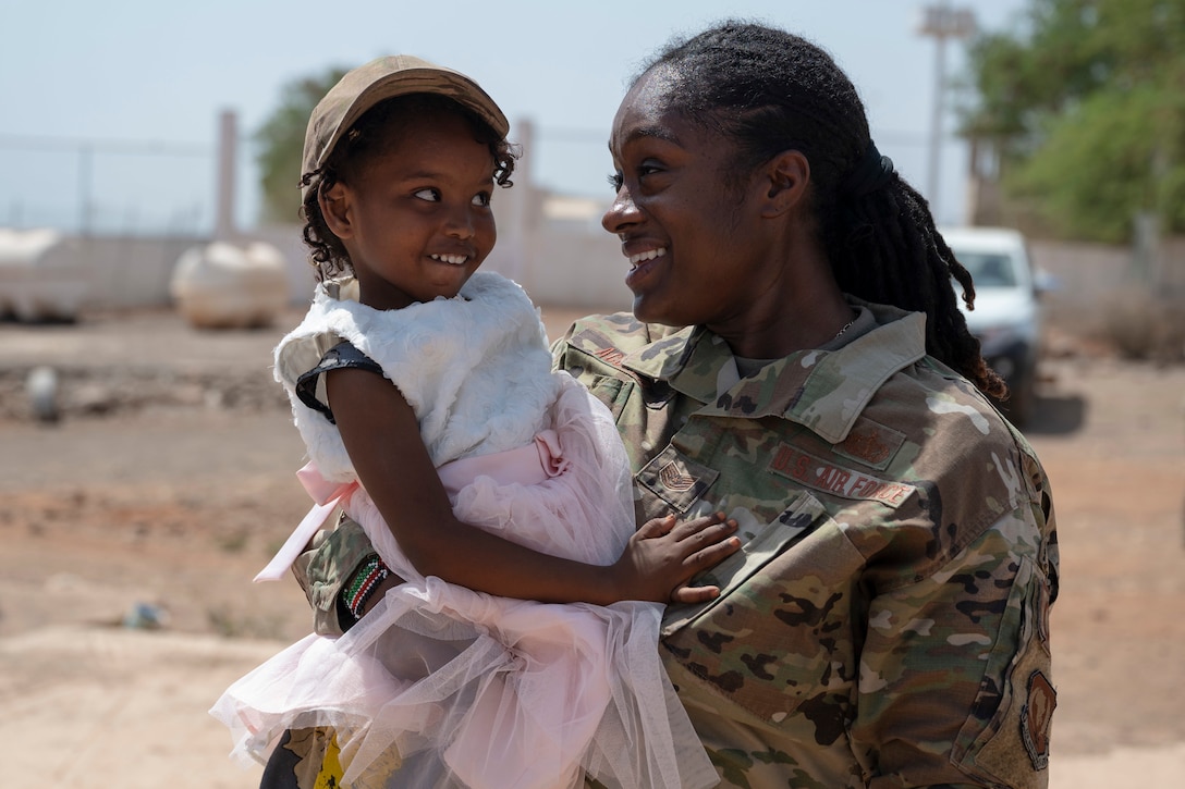 An airman holds a young girl wearing the soldier's hat as they smile at each other.