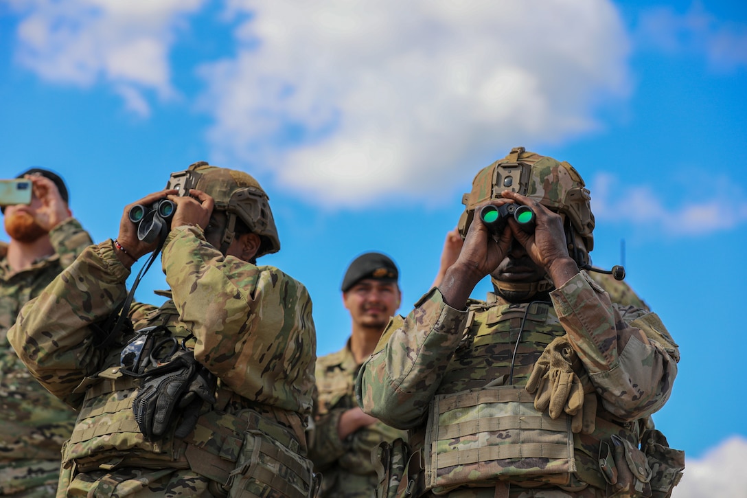 Two soldiers look through binoculars as two soldiers stand behind them.