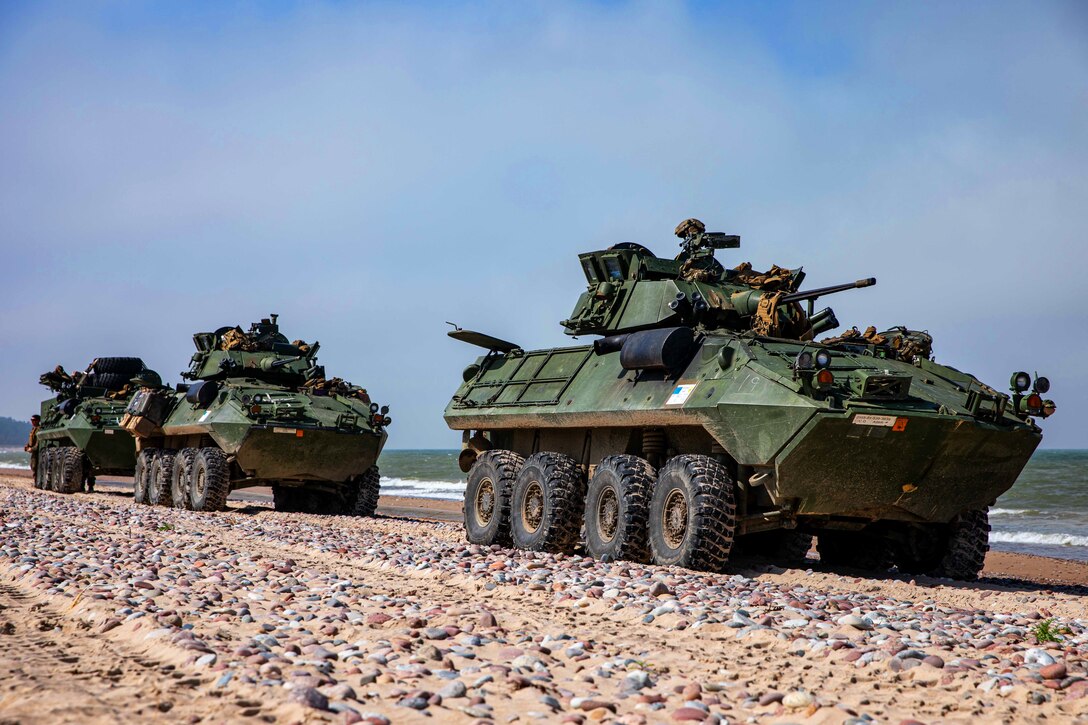 Three military vehicles drive along a stone-covered shoreline.
