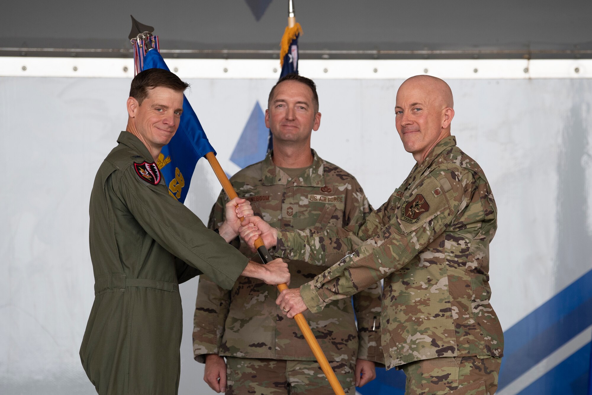 A photo of three men standing on a stage and "passing" a guidon from one to another.