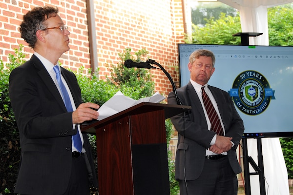 A man stands behind a lectern.  Another man stands near a television screen that displays the logo for the Defense Security Cooperation Agency.