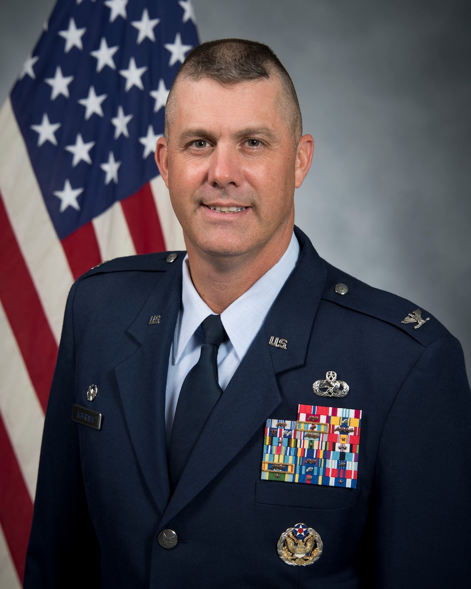 White male with brown hair poses in military dress uniform in front of American flag