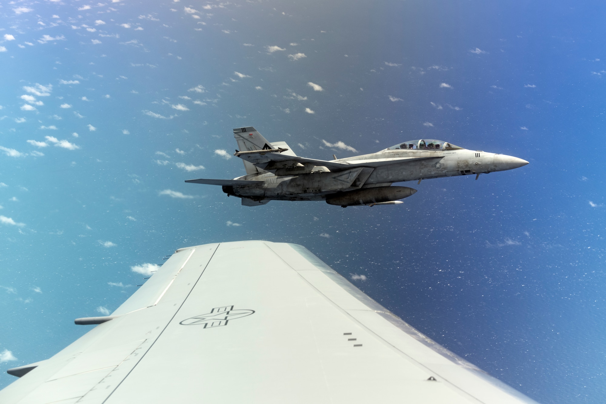 A U.S. Navy F/A-18 Hornet banks left in formation alongside a McConnell Air Force Base, Kansas, KC-46A Pegasus June 6, 2022, during Air Mobility Command’s Employment Concept Exercise 22-06. The F/A-18 just received fuel and is waiting for his wingman to finish his refuel by the Pegasus. McConnell aircrew and tankers provided aerial refueling in the Indo-Pacific Theater to practice joint interoperability in a multi-domain environment to build real-world proficiency and readiness. (U.S. Air Force photo by Master Sgt. John Gordinier)