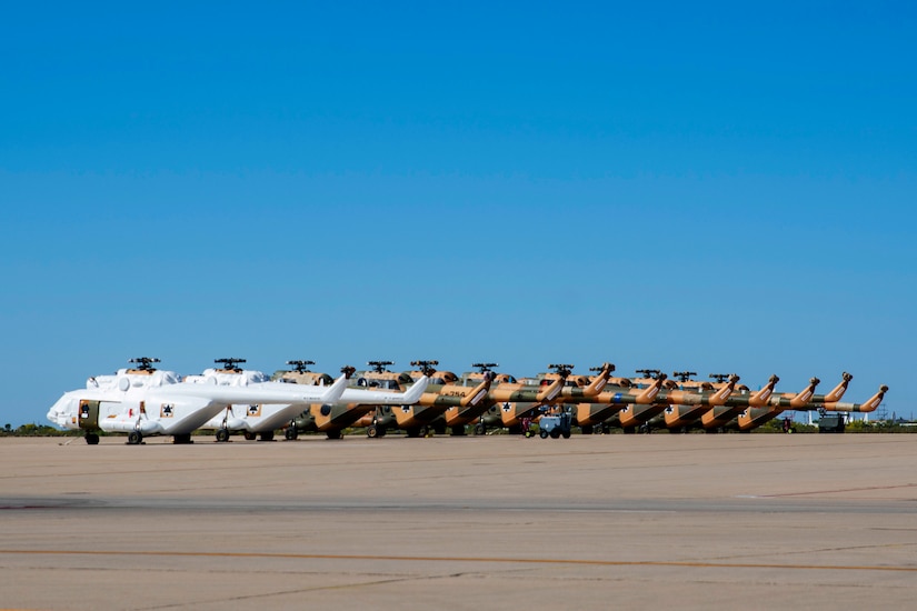 Two white helicopters and nine helicopters with camouflage paint schemes are lined on an airfield.