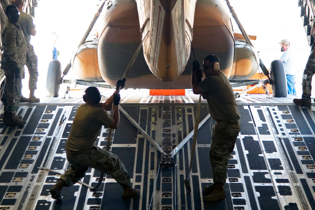 Two service members pull straps around the tail boom of a helicopter as it is loaded tail-first into the cargo hold of an aircraft.
