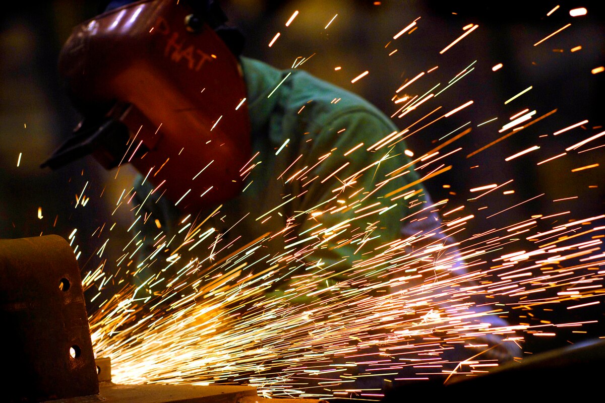 Sparks fly off of metalwork. A worker wears a protective hood.