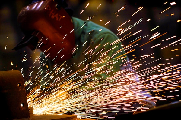 Sparks fly off of metalwork. A worker wears a protective hood.