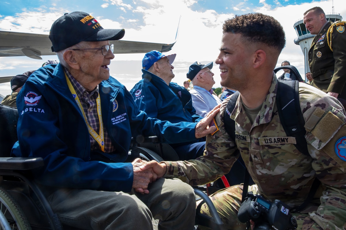 A smiling soldier crouches and shakes hands with a seated veteran amid a crowd of people outside.