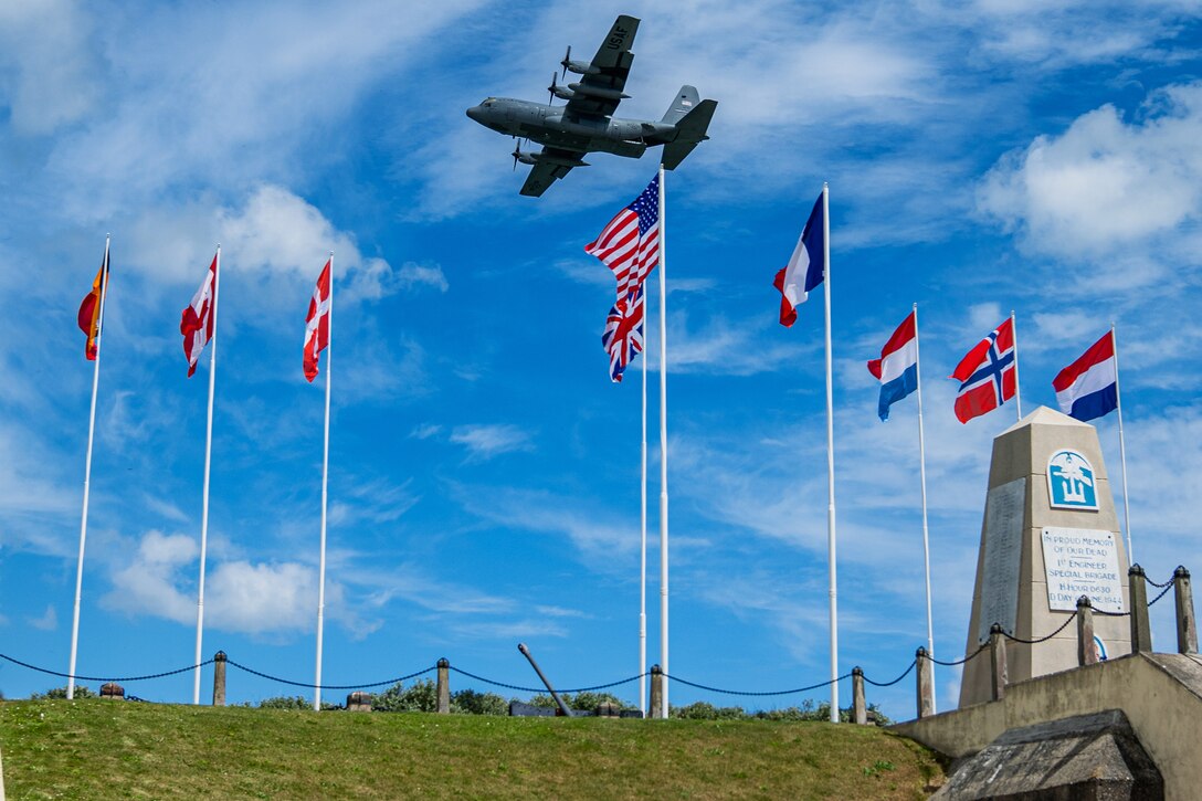 An aircraft flies over flags of multiple nations and a memorial at a grassy shoreline site.