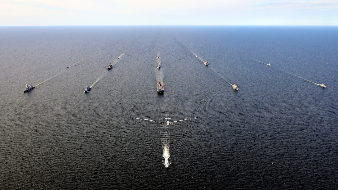 Ships sail in formation with aircraft flying in formation above them.