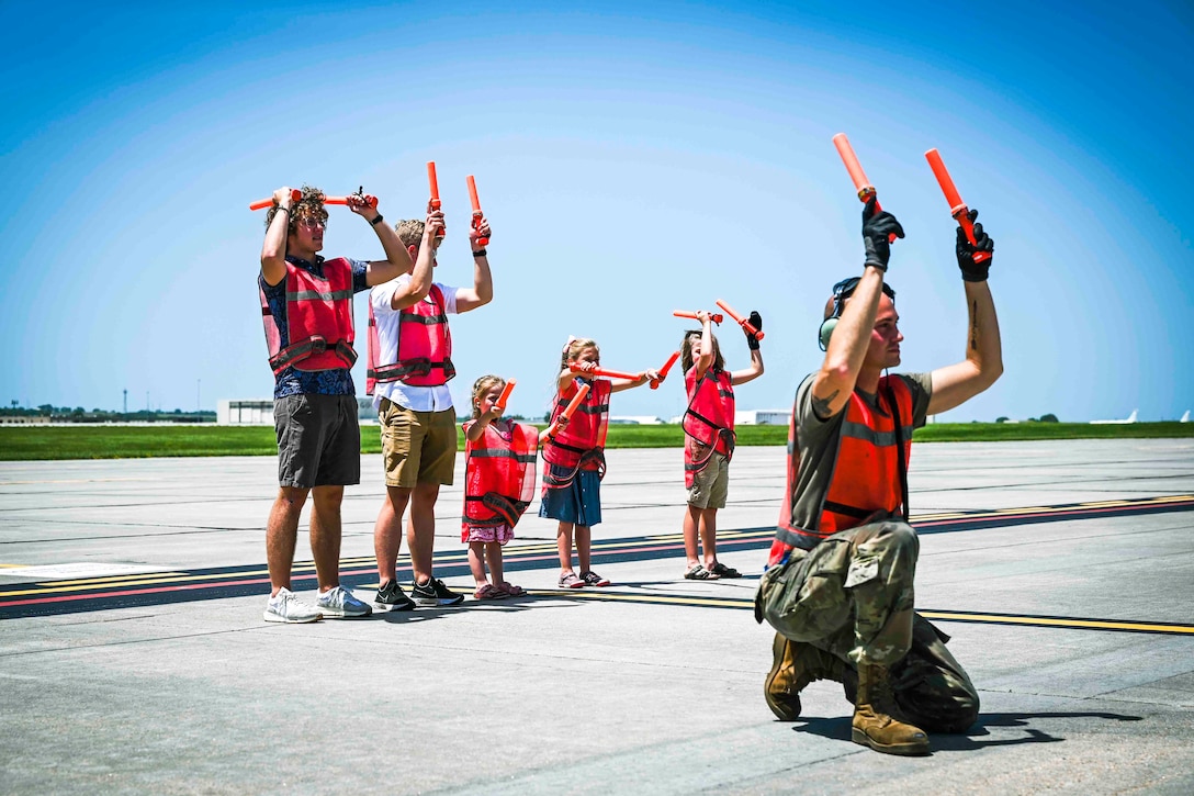 A group of people wearing red vests use wands to signal toward an aircraft, not pictured.