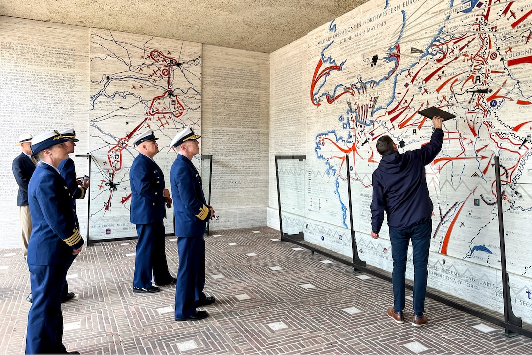 Coast Guardsmen stand in a room looking at a giant wall map as someone gestures toward it.