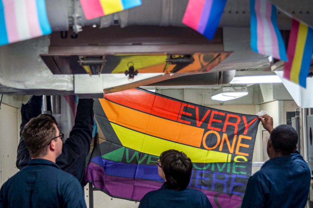 Sailors put up a rainbow flag in an interior area on a ship.