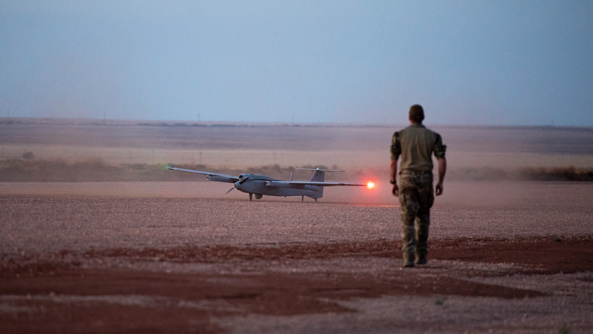 An Airman walks towards a small drone sitting on a dirt runway.