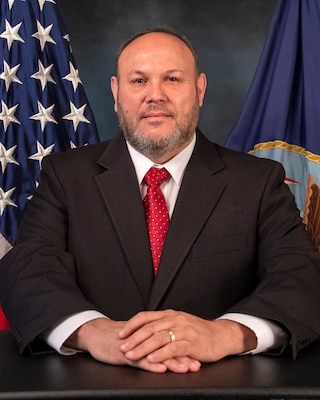 photo of Carlos A. Muñoz sitting in front of United States and Navy flags.