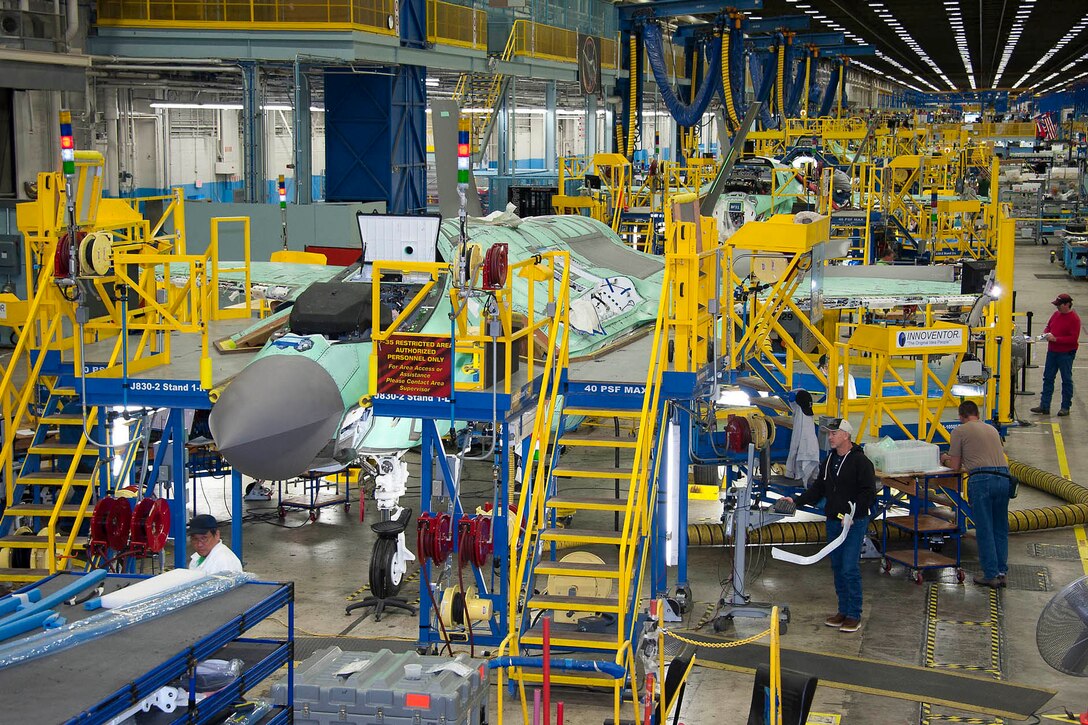In a large, brightly-lit facility with blue- and yellow-colored scaffolding, military aircraft are lined up in varying states of assembly.