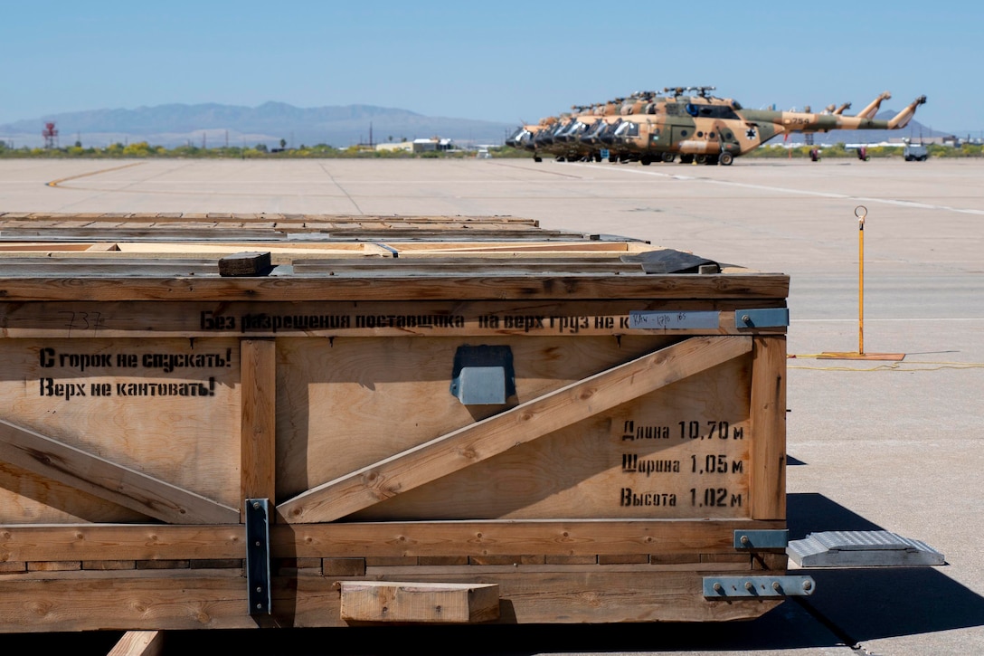 Several wooden crates sit in front several military helicopters on the flight line.
