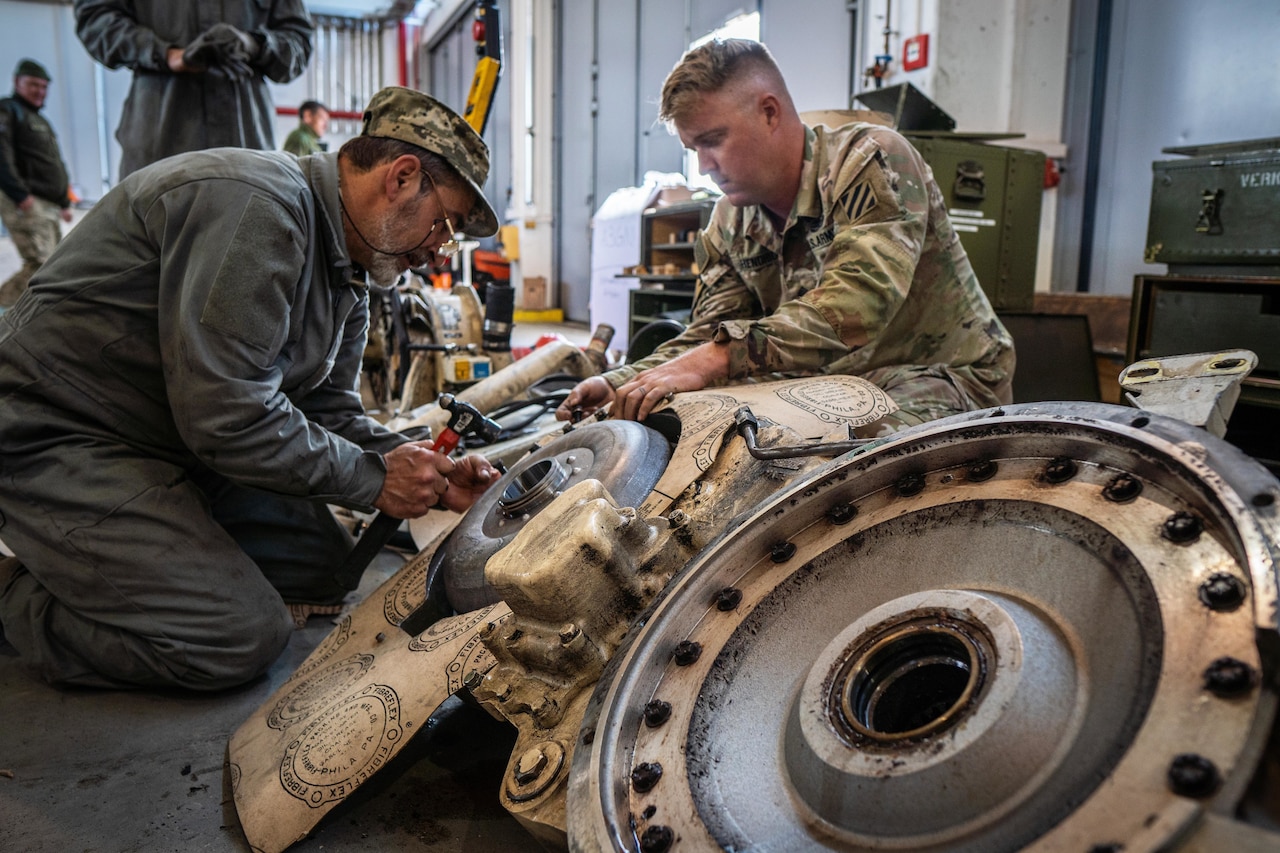 Two men, one wearing a military uniform, perform maintenance on a piece of equipment inside a building.