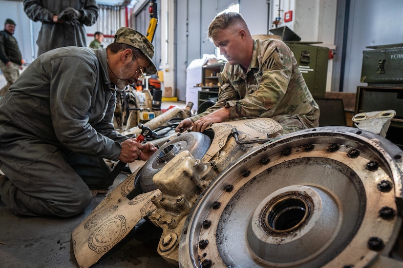 Two men, one wearing a military uniform, perform maintenance on a piece of equipment inside a building.