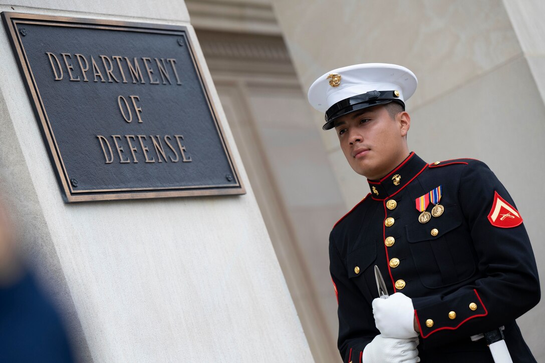 A Marine stands by a metal sign that says "DEPARTMENT OF DEFENSE" on an exterior Pentagon column.