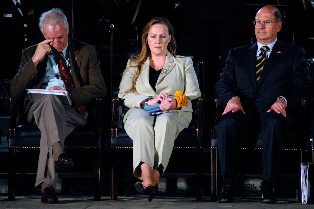 Three people sit beside each other. A woman in the middle holds a folded flag.