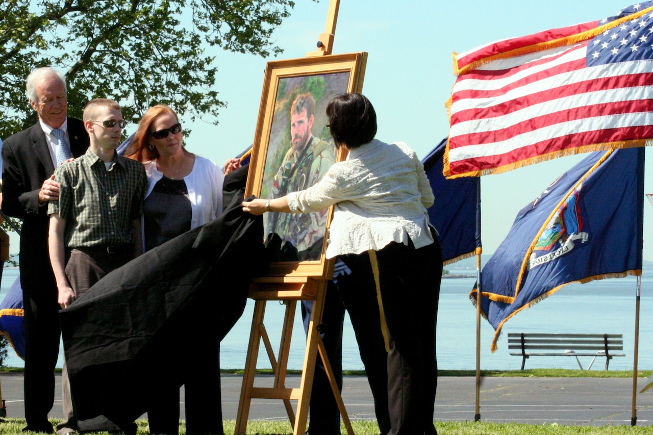 Three people look at a portrait of a man on an easel that’s being revealed by a woman.