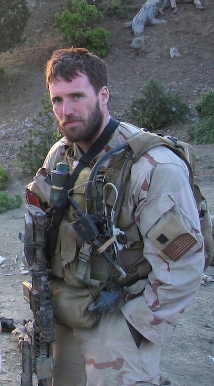 A man in combat gear looks at the camera while standing on a mountain.